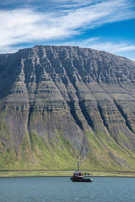 A scenic landscape photo of a large, layered mountain rising from a grassy slope, with a small boat floating on the water in the foreground under a blue sky with scattered clouds.