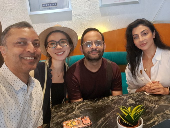 Four people pose together at a booth in a restaurant, smiling for a selfie during an Arts DAO event in Singapore. A sign reading "BREWERKZ" is visible on the wall behind them, and a small potted snake plant sits on the marble table alongside a phone.