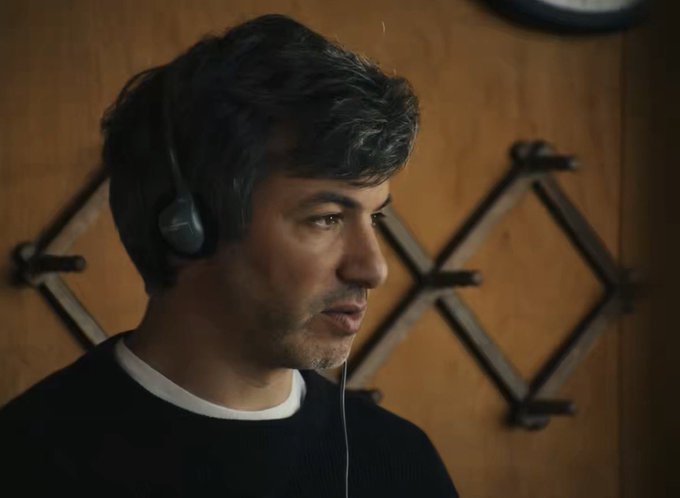 A man wearing a headset with a microphone, dressed in a black shirt, looks off to the side against a wooden background with decorative metal diamond patterns.