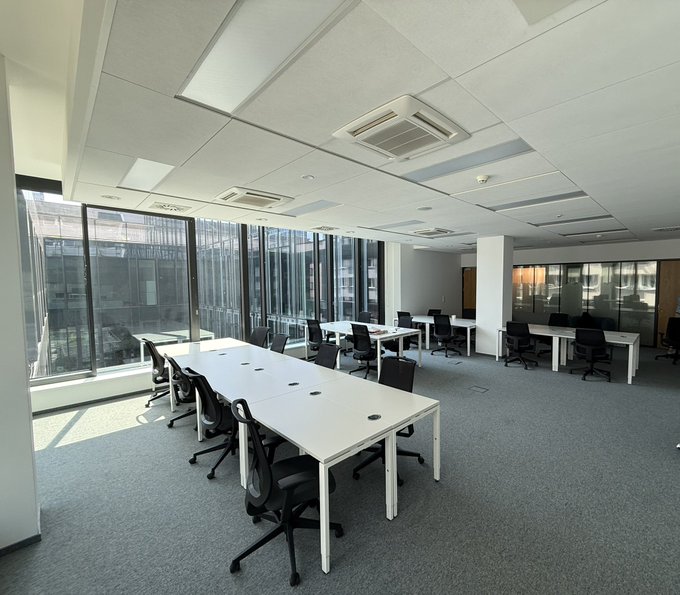 An empty, sunlit coworking space in Warsaw features rows of white desks paired with black ergonomic office chairs, set against floor-to-ceiling windows that flood the room with natural light. The minimalist setup, complete with modern ceiling lighting and gray carpeting, reflects the type of professional environment a crypto professional would look for in a shared office space.