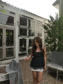 A young woman with long brown hair wearing a dark blue polka-dot dress stands on a gravel patio in front of a glass-paneled building.