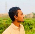 A short-haired young man in a yellow shirt stands in a field with an out-of-focus cityscape in the background.