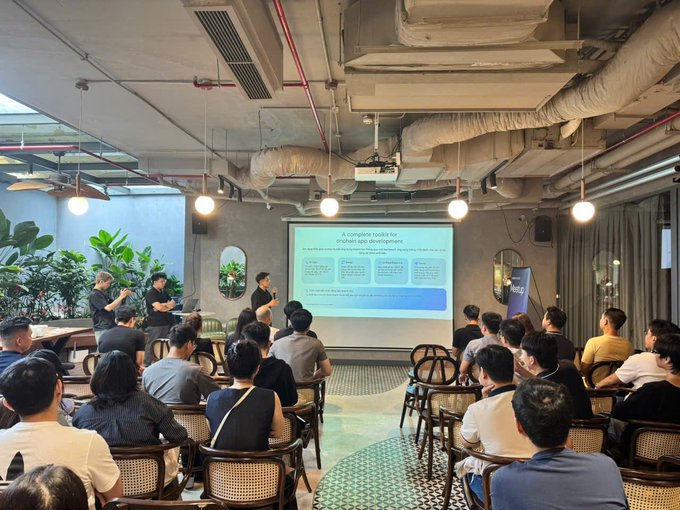 A presentation taking place in a modern industrial-style room, where a speaker addresses a seated audience in front of a screen displaying 'A complete toolkit for pitch and app development.'