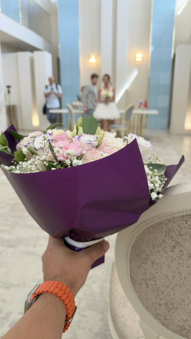 A first-person view of a hand holding a purple-wrapped bouquet of pink and white flowers, pointing toward a bride and groom in wedding attire at a reception venue.