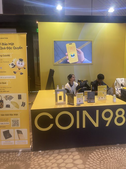 An event booth with yellow branding displaying "COIN98", featuring two staff members seated at the desk and a promotional banner advertising "Bảo Mật Quà Độc Quyền" with a cartoon mascot.