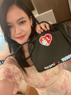 The image features a young woman holding up a black purse with stickers of cartoon characters, including a heart-shaped one. The purse has the words "Team1 x Pudgy Penguins" written on it. The girl appears to be posing for a photo, and there is also text in the image that reads "@AvaxTeam1 @pudgyph @pudgypenguins @philippinebc_wk".