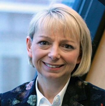 A woman with short blonde hair wearing a dark floral blazer over a white collared shirt smiles at the camera.
