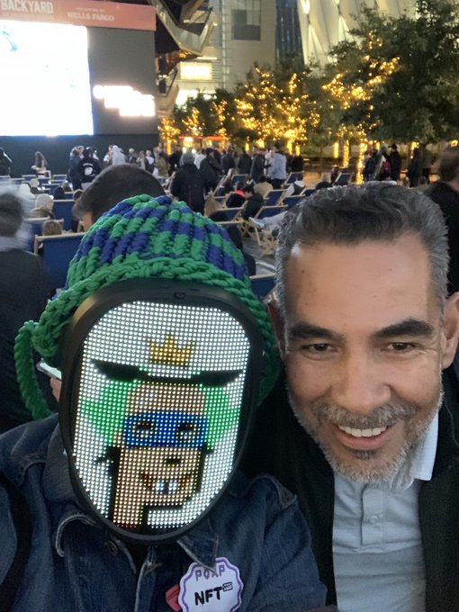 A man smiles beside someone wearing a green knit hat and an LED mask displaying a pixelated green-haired CryptoPunk, with an "NFT" button visible on the mask wearer's jacket and "BACKYARD WELLS FARGO" signage in the background of what appears to be an outdoor event. The playful image pairs perfectly with the post's message about embracing quirks and bringing your weird to crypto gatherings.