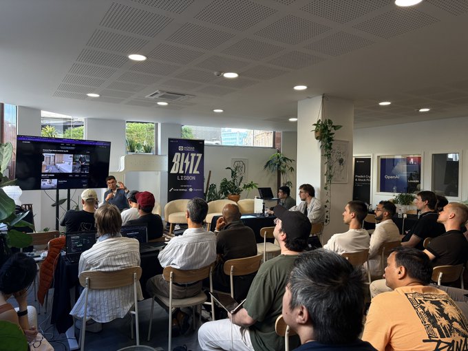 The image shows a group of people sitting in chairs, watching a presentation or demonstration taking place at a conference room. The attendees are engaged and focused on the event happening before them. There are several potted plants placed around the room, adding some greenery to the space. A TV screen is visible in the background, likely displaying the presentation being presented by @ivan_nomadz. The image also contains text that reads "full house tonight at LunarHQ for the @monad Blitz hackathon in Lisbon. got here just in time to catch a demo from @ivan_ Nomadz showcasing interoperability with solana liquidity." This suggests that the event is well-attended and of interest to those present, as it may be related to blockchain technology or other innovative projects.