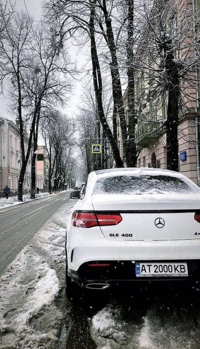 The image shows a white Mercedes-Benz car parked on a snowy street with snow falling from the sky. The car appears to be covered in snow, indicating that it has been parked there for some time. The scene also includes several other cars parked nearby, as well as trees lining the street and buildings in the background.