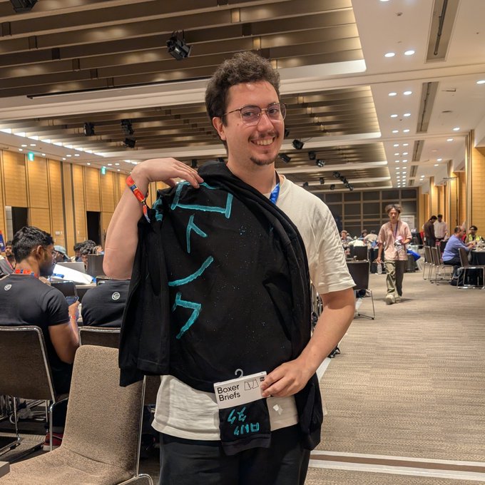 A smiling man with glasses and curly hair poses at what appears to be a convention, proudly holding up a black jacket with teal designs and a tag labeled 'Boxer Briefs L/XL'.