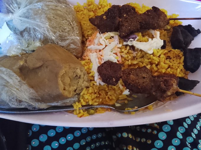 A plate of food featuring yellow rice topped with coleslaw, grilled meat skewers, what appears to be bread, and a spoon resting on the dish.