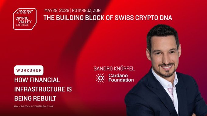 The image features a man wearing a suit and tie standing next to a red banner with white text that says "The Building Block of Swiss Crypto DNA" and "How Financial Infrastructure Is Being Rebuilt." The man appears to be smiling, possibly at the end of a workshop or presentation. The background is blurred, but it seems to be a conference room setting.