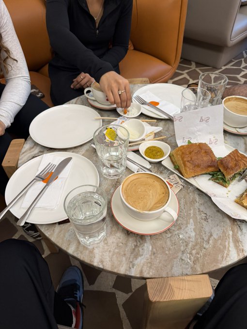 A dining table is set with empty white plates, silverware, two cups of coffee, water glasses, and a plate of sandwiches, with a person visible in the background.