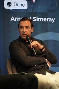 A man identified by the backdrop text as Arnaud Simeray speaks into a microphone during a Longitude panel discussion, seated in a black shirt and light pants. The image aligns with the post, which highlights his commentary on traditional banks facing competition from crypto platforms like Coinbase Prime for wealthy clients.