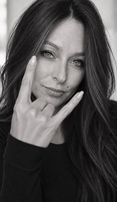 A black-and-white portrait of a woman with long dark hair making a peace sign with her hand, gazing directly at the camera.