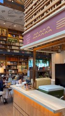 The image shows a cozy coffee shop with wooden walls, tables, chairs, and shelves filled with books. There are people sitting at tables enjoying their time, some working on laptops while others read or converse. The atmosphere appears inviting and comfortable, making it an ideal place for patrons to relax and engage in various activities such as reading, studying, or socializing.