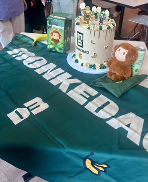 The image captures a celebratory table display featuring a green "MONKEDAQ" tablecloth, a cake adorned with DQ branding and figurines, plus stuffed animal mascots including a green lion and brown monkey. This setup visually reflects the 5th anniversary celebration of Monke, hosted by Superteam, with the DQ (Dairy Queen) branding complementing the festive milestone event.
