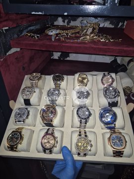 A display tray holding approximately sixteen assorted wristwatches in various styles and colors, with a gloved hand visible and additional watches/jewelry on a red surface in the background.