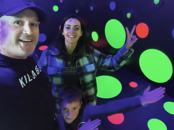 A family takes a selfie in a dark room illuminated with neon blacklight, with a man in a 'K7.8.6.1' shirt, a woman in plaid, and a young child surrounded by glowing green and red dots.