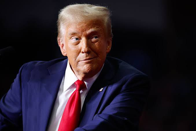 A portrait of Donald Trump wearing a navy blue suit with a red tie, looking to the side with a subtle expression against a dark background.