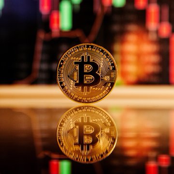 A physical gold Bitcoin coin stands prominently in focus against a blurred backdrop of a cryptocurrency trading chart, with its reflection mirrored on a glossy surface below. The image visually represents the significant $378.79 million Bitcoin whale transfer, symbolizing the high-value movement of 4,500 BTC from a dormant wallet.