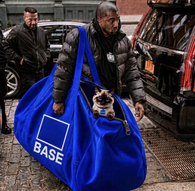 A man wearing a black puffer jacket carries a large blue "BASE" branded duffel bag containing a small cat wearing a blue collar, walking near luxury vehicles on a city street.