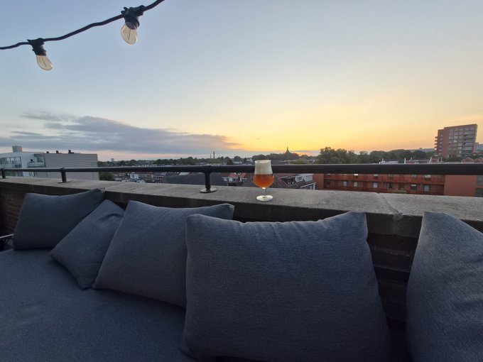 A rooftop terrace at sunset with string lights and gray cushions, featuring a glass of beer on the ledge overlooking the city skyline.