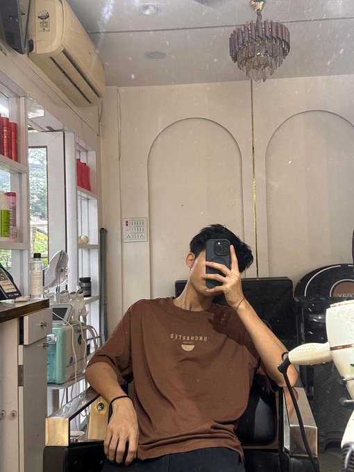 A person wearing a brown t-shirt takes a mirror selfie in a hair salon, holding a smartphone in front of their face.