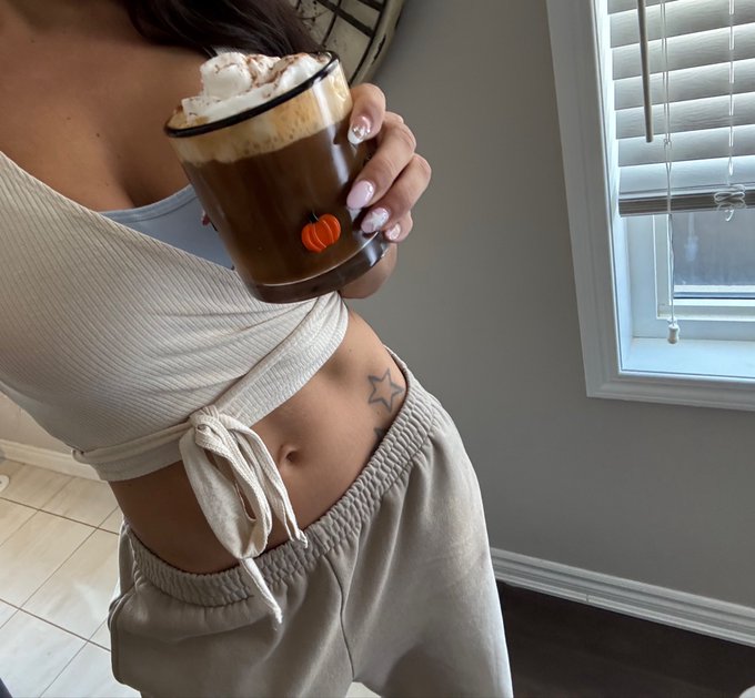 Photo of a woman holding a pumpkin-spiced latte drink, wearing a tied crop top and sweatpants, showing her midriff with a star tattoo.