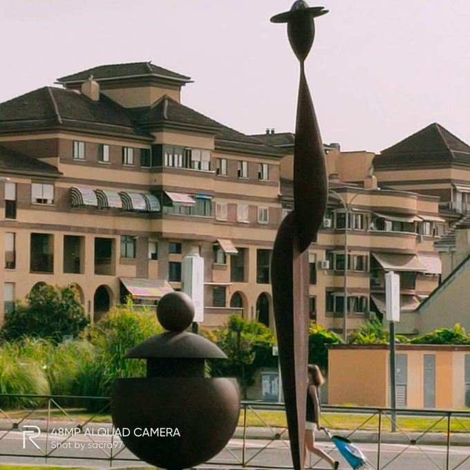The image shows two bronze sculptures—one tall, elongated figure with a disc-like head and a rounded, seated figure—set against a backdrop of a residential building, with a watermark reading "48MP AI QUAD CAMERA Shot by sacra97." In the context of the post asking about the sculptures in Leganés, the figures likely represent symbolic or cultural characters tied to the local community or artist commission.