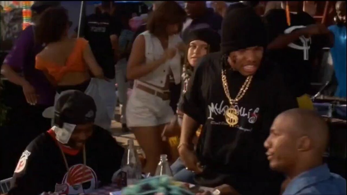 The image shows a group of people at an event, with one man wearing a black t-shirt that has "LOAN be like" written on it. The scene takes place around a dining table where several individuals are gathered, including a person sitting in front of the table and others standing nearby. There is also another person seated further back from the main group.