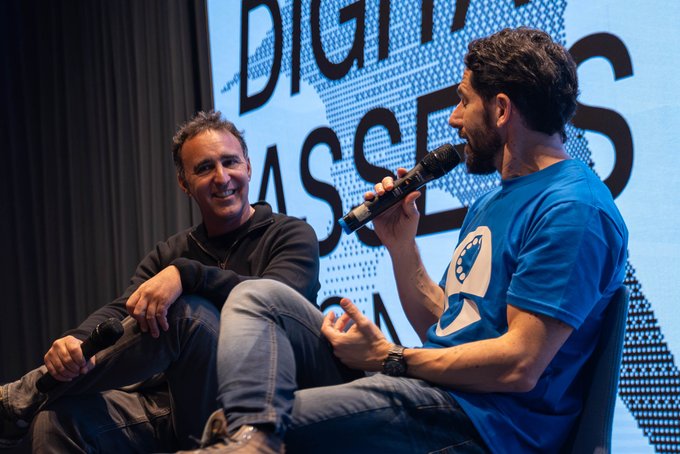 Two men seated on a stage at a 'Digital Assets' event, with one man in a blue shirt holding a microphone and speaking while the other smiles, holding a phone.