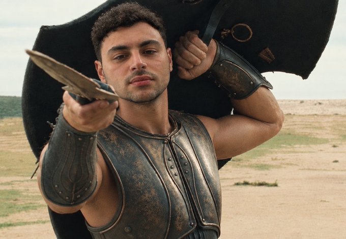 A man in ancient Greek or Roman-style bronze armor poses with a spear held forward, carrying a large dark shield on his back, in a desert-like setting.