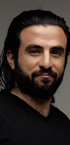 A portrait photograph of a man with dark hair and a full beard, wearing a black shirt against a gray background.
