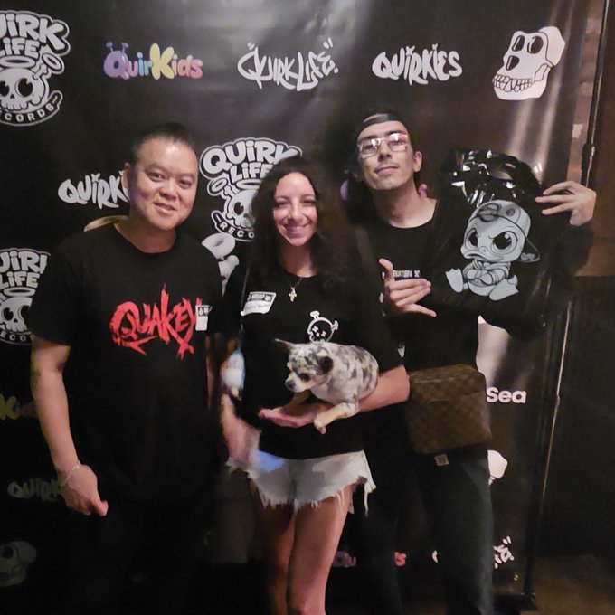 Three people pose in front of a 'Quirk Life' branded backdrop alongside a woman holding a small dog, with various brand logos visible including 'Quirkids', 'Quinkies', and 'Quirk Life Records'.