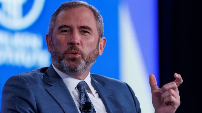 A bearded man in a navy blue suit and dark tie is speaking at a formal event, gesturing with his right hand as he makes a point. Behind him, a blue backdrop displays partial text reading "FERENCE" and "N FRANC," suggesting a conference setting in San Francisco. The image accompanies the post about Ripple CEO Brad Garlinghouse discussing how XRP offers faster and lower-risk cross-border payments compared to traditional systems like SWIFT.