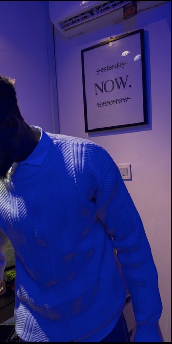 A person in a blue sweater leans forward in a purple-lit room beneath a framed print reading 'yesterday NOW. tomorrow'.