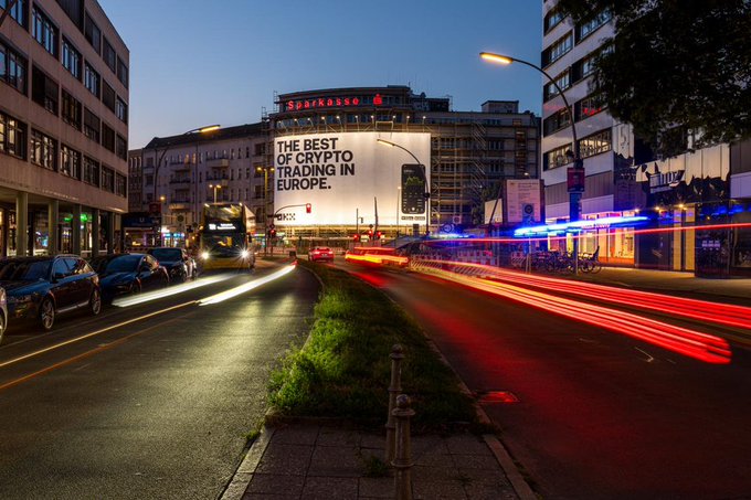 A long-exposure nighttime shot of a Berlin street reveals a prominent illuminated billboard displaying the OKX advertisement "THE BEST OF CRYPTO TRADING IN EUROPE" alongside the iconic red Sparkasse sign atop the building. Streaking red and white light trails from passing vehicles emphasize the dynamic urban setting, reinforcing the post's theme of impactful crypto advertising in the German capital.