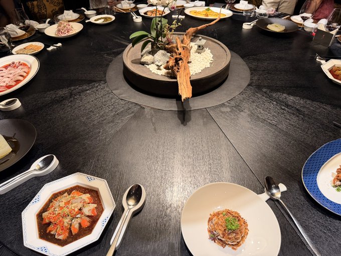 A round dining table set for a meal, featuring a central decorative platter with white pebbles and a wooden sculpture, surrounded by various Asian dishes, including what appears to be mapo tofu and noodles.