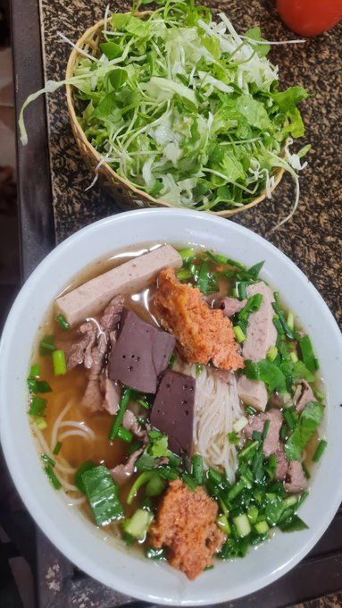 A bowl of Vietnamese noodle soup containing rice noodles, beef slices, congealed blood cubes, fried tofu, pork, and scallions in broth, served alongside a basket of fresh herbs and greens.
