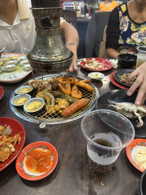Photograph of a Japanese-style meal featuring a tabletop grill with various seafood and meats, surrounded by small side dishes, sauces, and drinks on a wooden table.