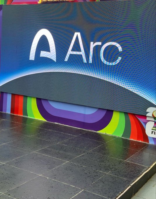 An 'Arc' logo is displayed on a large digital screen with a colorful rainbow wave design at the bottom, set against a tiled floor.