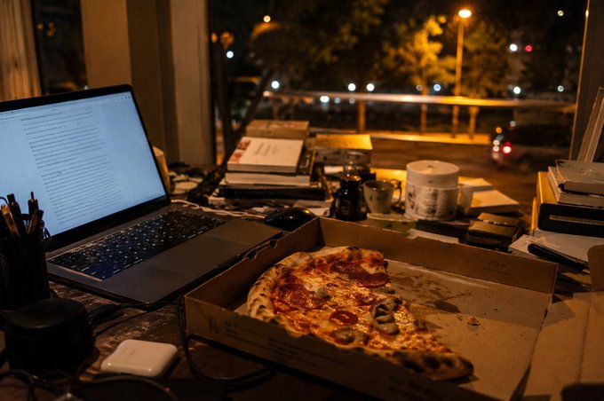 An atmospheric nighttime photo of a desk by a window, featuring an open MacBook laptop displaying text, an open pizza box with a half-eaten pepperoni and mushroom pizza, coffee cups, books, and city lights visible through the window.