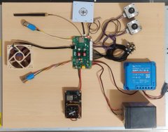 An electronics project setup on a wooden surface showing a circuit board connected to various components including a cooling fan, motors, a blue battery charger, and a small digital display.