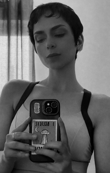 A black-and-white mirror selfie of a person with short dark hair taking a photo with a smartphone that has an 'I BELIEVE' UFO card in a wallet case.