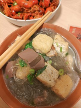 A bowl of Chinese soup containing glass noodles, tofu puffs, congealed duck blood, and pork blood, garnished with green onions, with wooden chopsticks resting on top and a bowl of crawfish visible in the background.