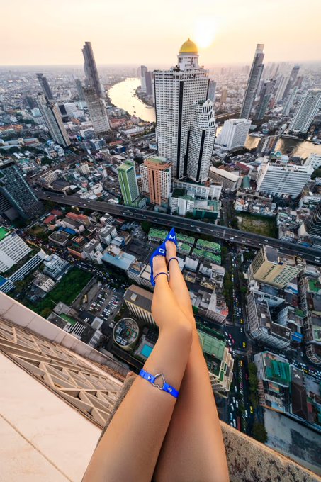 Two manicured hands adorned with diamond rings extend from a rooftop ledge high above Bangkok, with the State Tower and Chao Phraya River visible below at sunset. The image showcases Angela Nikolau's signature style of rooftop engagement photography, as referenced in her "Memes" NFT collection Card 92, "Gm From a Roof."
