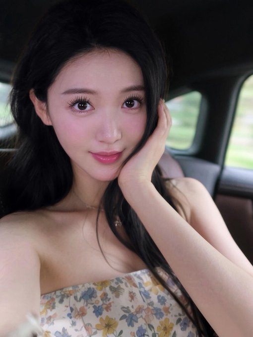 A young woman with long dark hair and pink blush rests her face against her hand, wearing a strapless floral top inside a vehicle.
