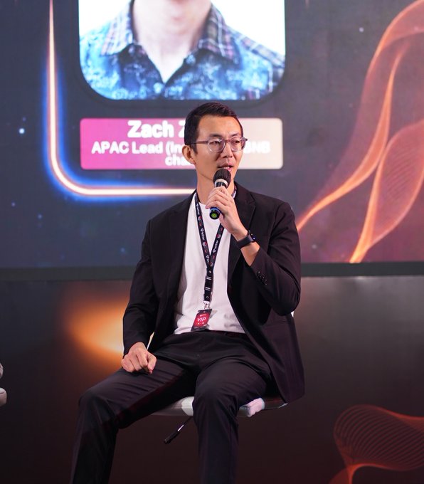 A man in a black suit and glasses speaks into a microphone while seated on stage, with a screen behind him displaying "Zach Z... APAC Lead" and a plaid-shirt avatar. The image appears to capture a moment from the BNB Chain booth at the Indonesia Blockchain Week event referenced in the post.